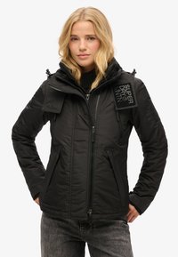 Veste noire en duvet avec plusieurs poches zippées, col haut, tissu texturé, capuche ajustable et un écusson logo sur l'épaule.
