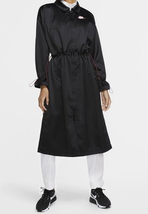 Osoba nosící černý kabát délky ke kolenům se staženým pasem, bílé kalhoty a černé tenisky Nike s bílými podrážkami a logy ve tvaru háčka.