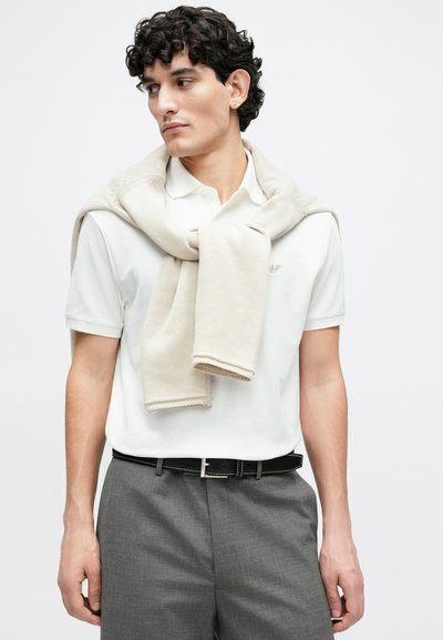 Béžový pletený sveter prehodený cez biele polo tričko, doplnený o sivé nohavice. Doplnky zahŕňajú čierny opasok so striebornými kovaniami.