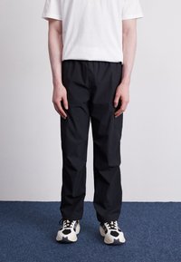 Person som står i en vit t-shirt, svarta byxor och vita och svarta sneakers på en blå matta mot en enkel vit vägg.