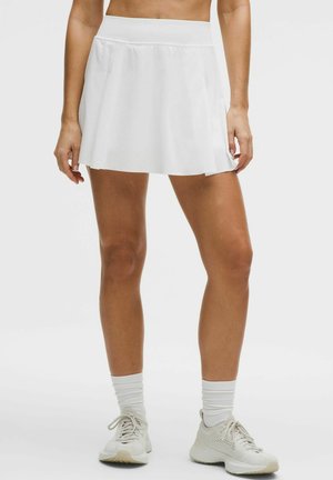 Mujer vestida con una falda deportiva blanca, calcetines blancos y zapatillas blancas, de pie contra un fondo claro y liso.