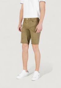 Pantaloni corti verde oliva con finitura testurizzata, dotati di tasche frontali angolate, bottone anteriore e vestibilità slim. Abbinati a sneaker bianche.