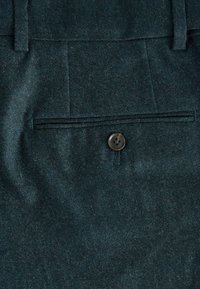 Ciemny teal wełniany materiał o gładkiej teksturze. Posiada pojedyncze zapięcie na guzik oraz tylną kieszeń z delikatnymi fałdami.