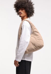 Borsa a spalla imbottita color sabbia con una consistenza morbida, forma arrotondata e una grande apertura. Presenta un logo sottile impresso sul lato.