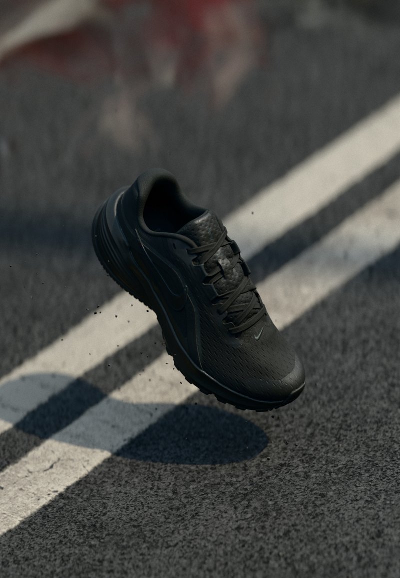 Chaussure de course noire en l'air au-dessus d'une route asphaltée avec des marquages blancs, petits débris visibles près de la chaussure.