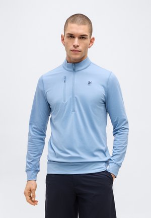 Homme aux cheveux courts portant un pull léger bleu clair à fermeture éclair partielle et un pantalon sombre, debout devant un fond clair uni.