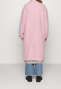 Langer, übergroßer pinker Mantel aus weichem Stoff, mit geknöpften Manschetten und schlitz am Rücken, kombiniert mit einem grauen Pullover und weitem blauen Jeanshose.