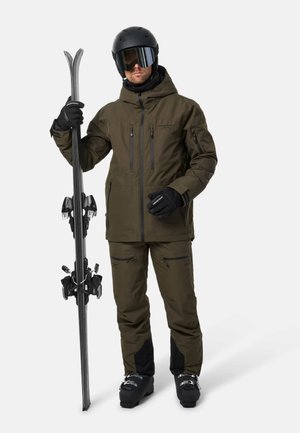 Un esquiador con chaqueta y pantalones de color verde oliva, casco, gafas y guantes sostiene los esquís en posición vertical sobre un fondo blanco liso.
