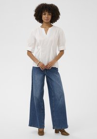 Top alb cu mâneci bufante și decolteu în V, asortat cu blugi bleu cu picioare largi și pantofi cu toc bloc maro. Design simplu și curat.