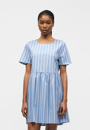 Donna con capelli intrecciati che indossa un vestito a maniche corte a righe verticali blu e bianche, in piedi davanti a uno sfondo bianco semplice.