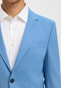 Uomo che indossa un blazer azzurro chiaro testurizzato sopra una camicia bianca con bottoni crema, mostrato dal collo fino a metà busto.