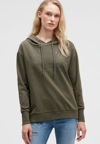 Olivgrüner Kapuzenpullover mit einer Fronttasche, Kordelzugkapuze und geprägtem Textdetail. Weiches Material und lässige Passform. Kombiniert mit hellblauen Jeans.