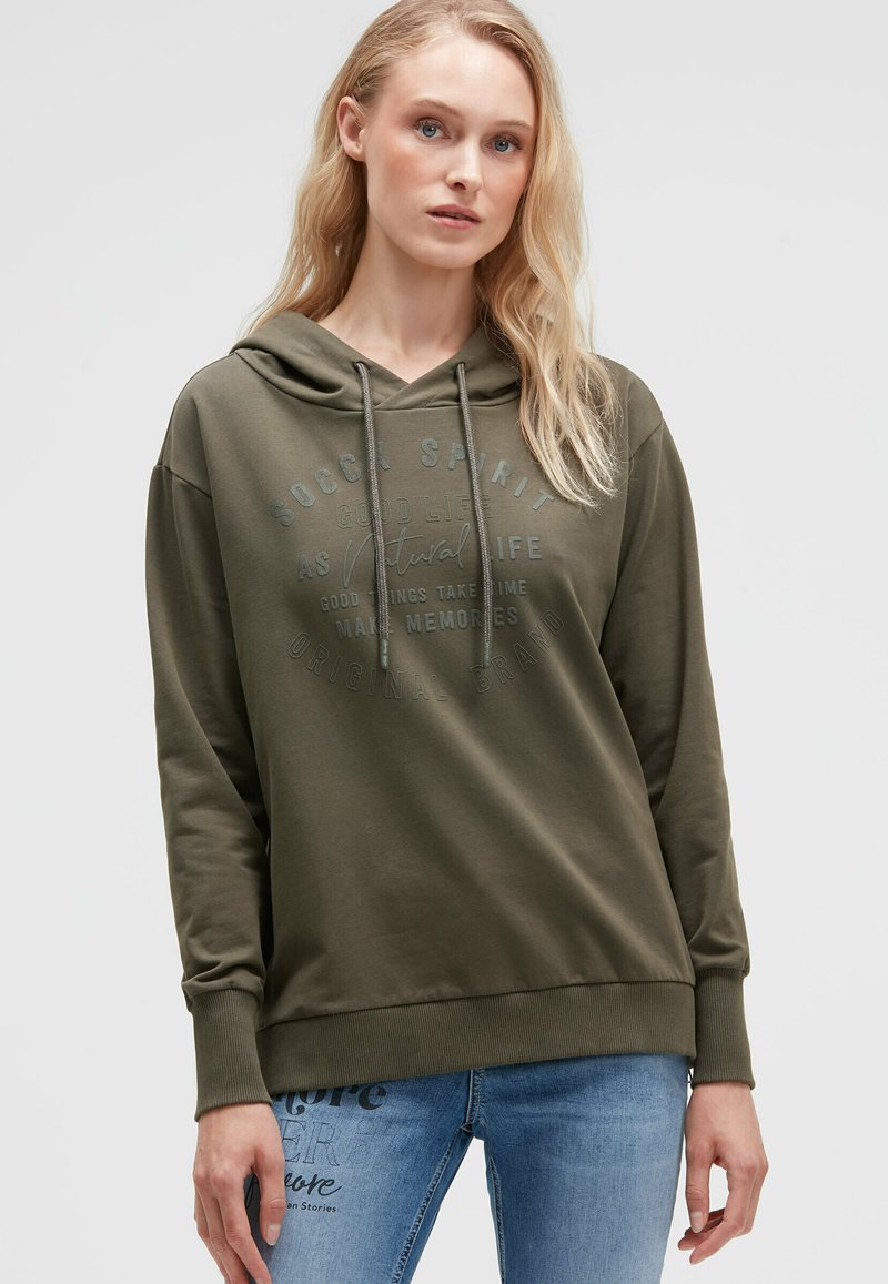 Olivgrüner Kapuzenpullover mit einer Fronttasche, Kordelzugkapuze und geprägtem Textdetail. Weiches Material und lässige Passform. Kombiniert mit hellblauen Jeans.