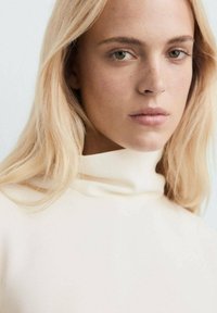 Maglione a collo alto di colore crema con una texture liscia. Il modello ha lunghi capelli biondi e lisci e un trucco naturale.