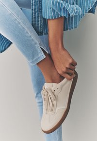 Pantof sport din piele bej, cu detalii perforate și șireturi crem. Asortat cu blugi bleu deschis, având un tiv despărțit la nivelul gleznei.