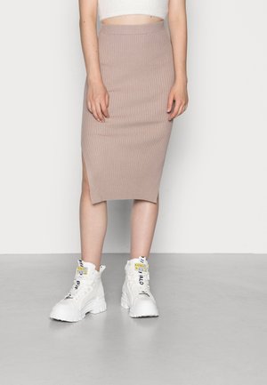 Kvinde iført en beige ribstrikket midinederdel med sideløsning og hvide chunky høje sneakers med sorte tekstdetaljer.