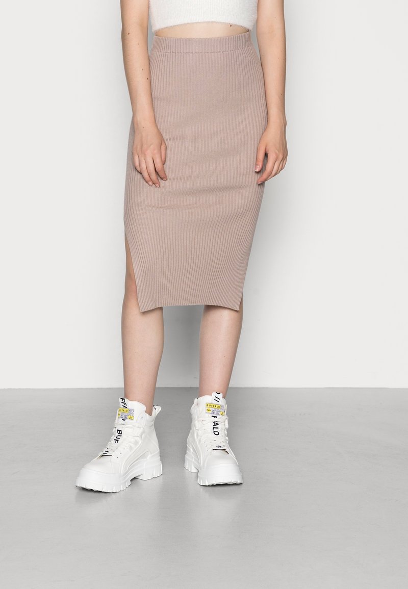 Femme portant une jupe midi en maille côtelée beige avec une fente latérale et des baskets montantes blanches épaisses avec des détails de texte noirs.
