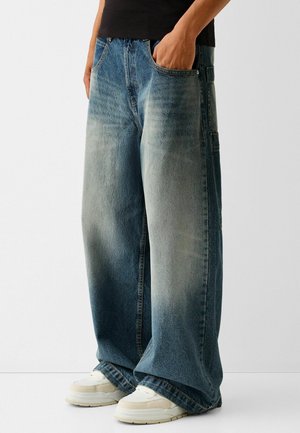 Personne portant un jean bleu délavé à jambes larges et des baskets blanches, main dans la poche, debout devant un fond uni.