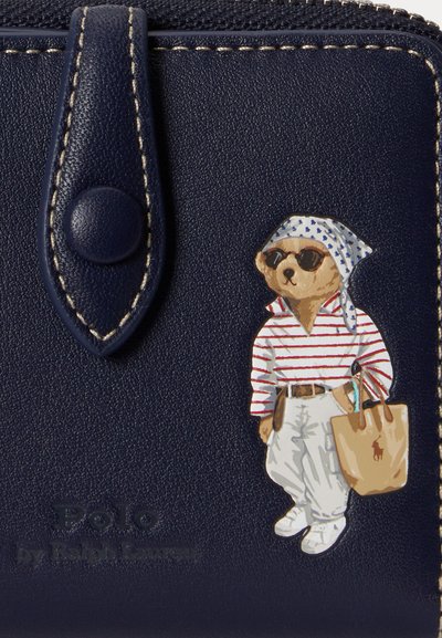 Portefeuille en cuir bleu marine avec bords cousus, présentant un ours portant des lunettes de soleil, une chemise rayée, un pantalon blanc, un foulard et portant un sac fourre-tout beige.