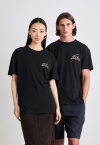 Tricourile negre din bumbac au un mic logo multicolor "JEANS" pe partea stângă a pieptului. Un model poartă o fustă din denim maro; celălalt, pantaloni scurți bleumarin.
