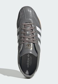 Γκρι δερμάτινο αθλητικό παπούτσι Adidas με λευκές ρίγες, κορδόνια και χρυσό λογότυπο της μάρκας στη γλώσσα, απεικονίζεται από πάνω σε φωτεινό φόντο.