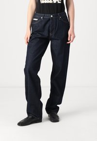 Vaqueros de mezclilla azul oscuro con un corte holgado, que presentan un diseño clásico de cinco bolsillos y costuras amarillas. Combinados con mocasines negros.