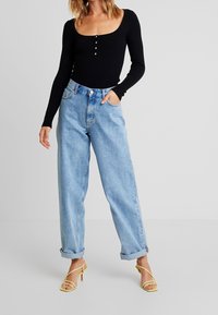 Femme portant un haut côtelé noir à manches longues, un jean bleu clair ample taille haute avec revers, et des sandales à talons jaunes à lanières, debout avec une main dans la poche.