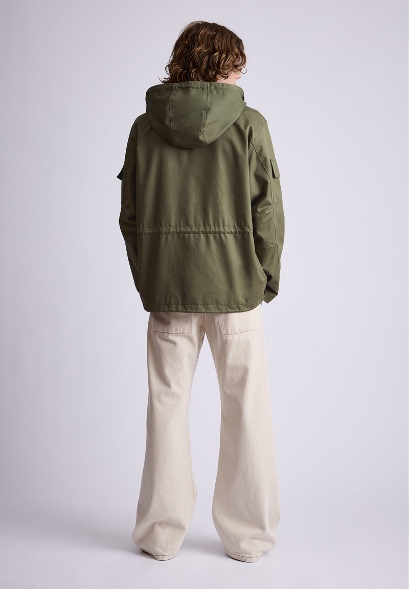 Personne aux cheveux bouclés de dos portant une veste à capuche verte oversize et un pantalon large crème sur un fond uni.