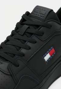 Sapatilha preta de couro com superfície texturizada, atacadores pretos, língua acolchoada e um logótipo em vermelho, branco e azul na lateral.