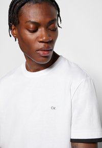 T-shirt bianco in cotone con scollatura rotonda, logo CK nero sulla parte sinistra del petto e maniche corte con bordi neri. Tessuto morbido.