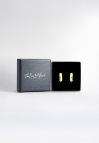 Guldtonade örhängen med en böjd design, dekorerade med små klara stenar, presenterade i en svart texturerad smyckeslåda med en sammetsinredning.