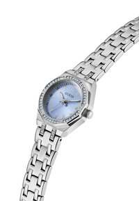 Montre Guess en argent avec un cadran bleu, des index horaires en chiffres romains, une lunette ornée de cristaux et un bracelet en métal à maillons.