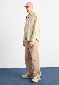 Světlobéžová košile na knoflíky, pod ní šedé tričko, hnědé volné kalhoty a růžová čepice. Na nohou béžovo-modré tenisky, stojí na modrém koberci.