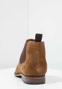 Stivale Chelsea in suede marrone con pannello elastico laterale scuro e linguetta, tacco in legno basso, fotografato su una superficie e sfondo bianchi.