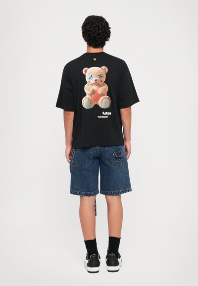 T-shirt nera a maniche corte con una grafiche di un orso colorato sulla schiena. Abbinata a pantaloni corti di jeans e calzini e scarpe da ginnastica nere.