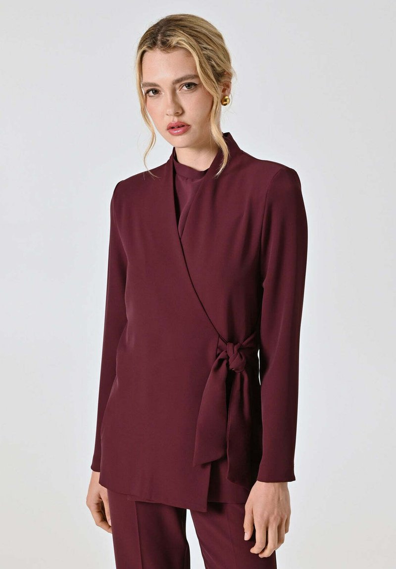Top avvolgente color bordeaux realizzato in tessuto liscio con maniche lunghe, una fusciacca in vita e un design con sovrapposizione leggera. Taglio semplice ed elegante.