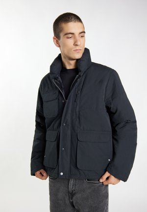 Junger Mann trägt eine schwarze gesteppte Jacke mit mehreren Fronttaschen und einem hohen Kragen, schaut leicht seitlich vor einem einfarbigen Hintergrund.