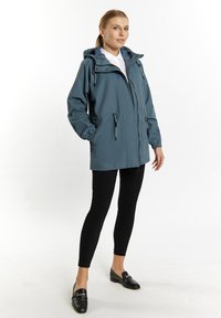 Vandafvisende blå jakke med hætte, snore og frontlynlås. Kombineret med sorte leggings og sorte loafers. Glat tekstur.