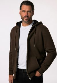 Brun zip-up hoodie med snoretræk i hætten, ribbede manchetter og forlommer. Fleecforing synlig ved kraven; båret over en hvid skjorte.