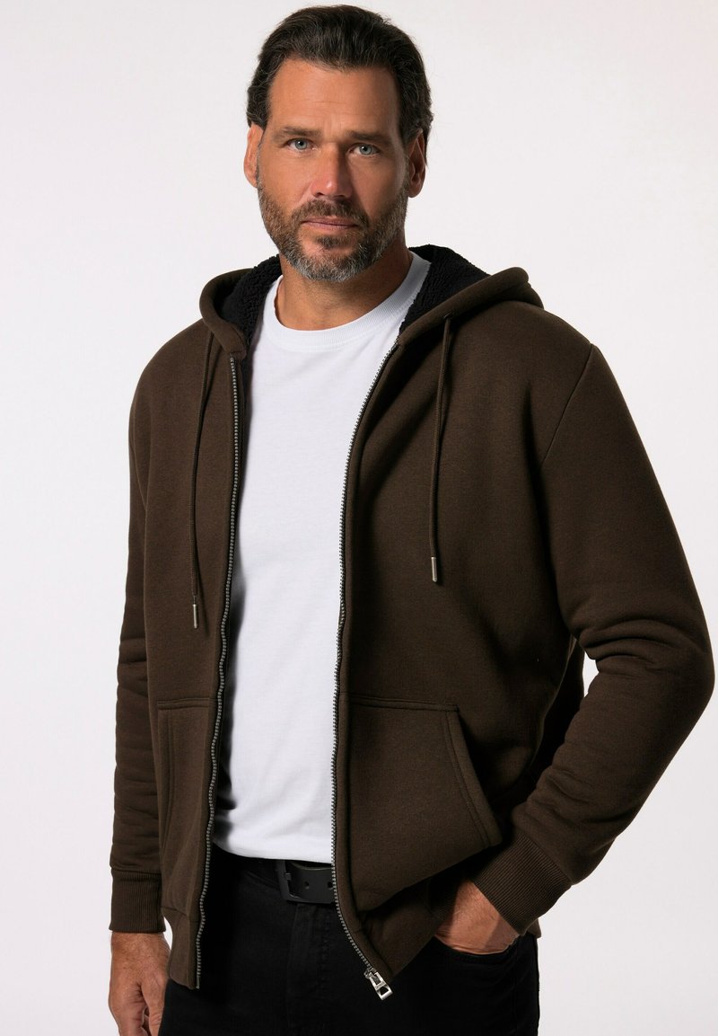 Brun zip-up hoodie med snoretræk i hætten, ribbede manchetter og forlommer. Fleecforing synlig ved kraven; båret over en hvid skjorte.