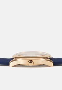 Montre ronde avec un boîtier en métal doré rose, un bracelet en silicone bleu et un verre plat. Présente une couronne texturée sur le côté droit.