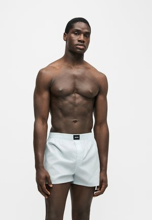 Homme musclé debout, posture détendue, portant un boxer gris clair avec une étiquette noire « BOSS » sur la ceinture, sur un fond uni.