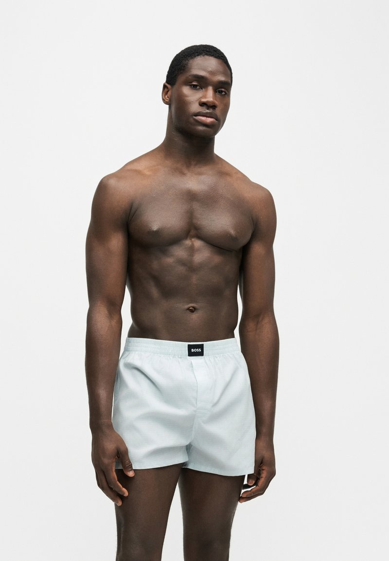 Homme musclé debout, posture détendue, portant un boxer gris clair avec une étiquette noire « BOSS » sur la ceinture, sur un fond uni.