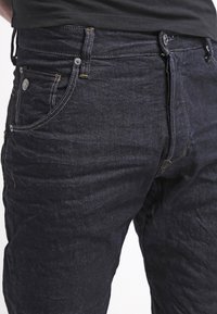 Unos vaqueros de mezclilla azul oscuro con cinco bolsillos, un cierre clásico de botón y cremallera, con acentos de costura amarilla y un acabado ligeramente texturizado.