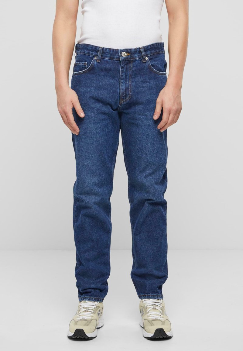 Jeans de mezclilla en azul oscuro con un diseño clásico de pierna recta, lazos para cinturón, bolsillos frontales y cremallera. Llevados con zapatillas deportivas en tonos neutros.