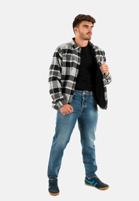 Camicia in flanella a quadri nera e bianca, sbottonata sopra una maglietta nera, abbinata a jeans blu e scarpe da ginnastica scure con suola in gomma.