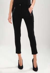 Schwarze Skinny-Jeans mit Reißverschluss-Seitentaschen, mittelhoher Taille und verkürzter Länge. Hergestellt aus elastischem Stoff mit glatter Textur.