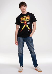 Schwarzes Baumwoll-T-Shirt mit einem bunten Iron Man-Design. Kombiniert mit blauen Jeans und lässigen weißen Schuhen mit hellorangefarbenen Sohlen.