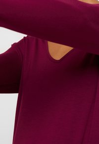 Langarmshirt in tiefem Burgunderrot mit V-Ausschnitt. Der Stoff wirkt glatt und elastisch, hat ein tailliertes Design und nahtlose Ärmel.