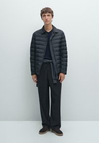 Schwarze Steppjacke mit horizontalen Nähten, marineblaues Hemd, grauer Pullover um die Taille gebunden, lockere dunkle Hose und dunkle Schuhe. Minimalistisches Design.